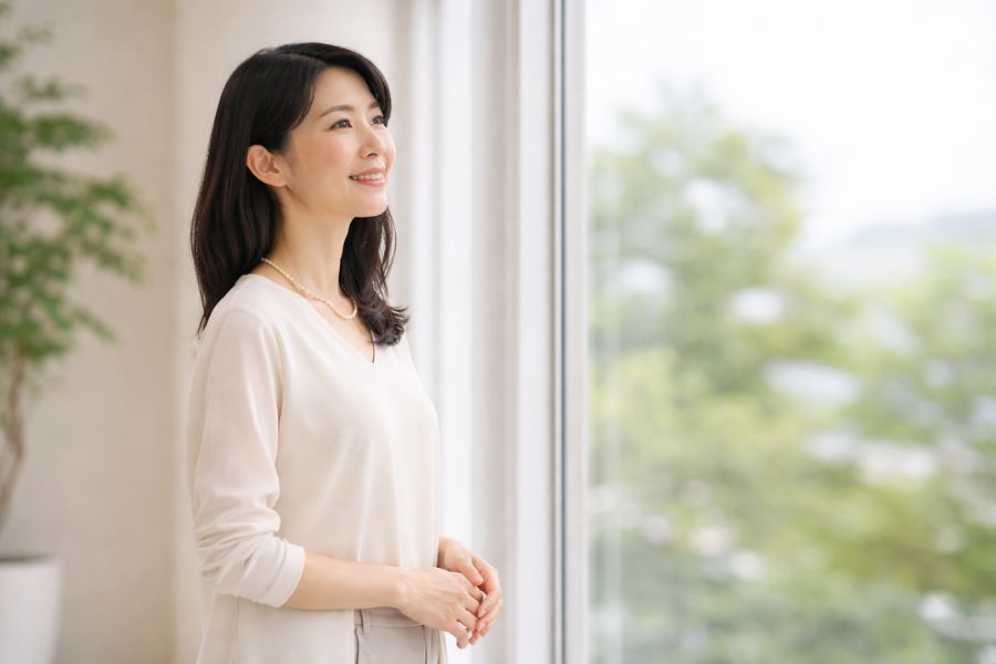 大きな窓の前に立ち外を見ながら微笑んでいる日本人女性