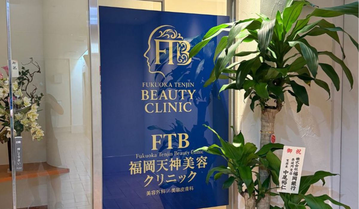 白衣に名札の若い日本人男性医師が美容クリニック入口前に立ち手を軽く差し出して招くワイドショット