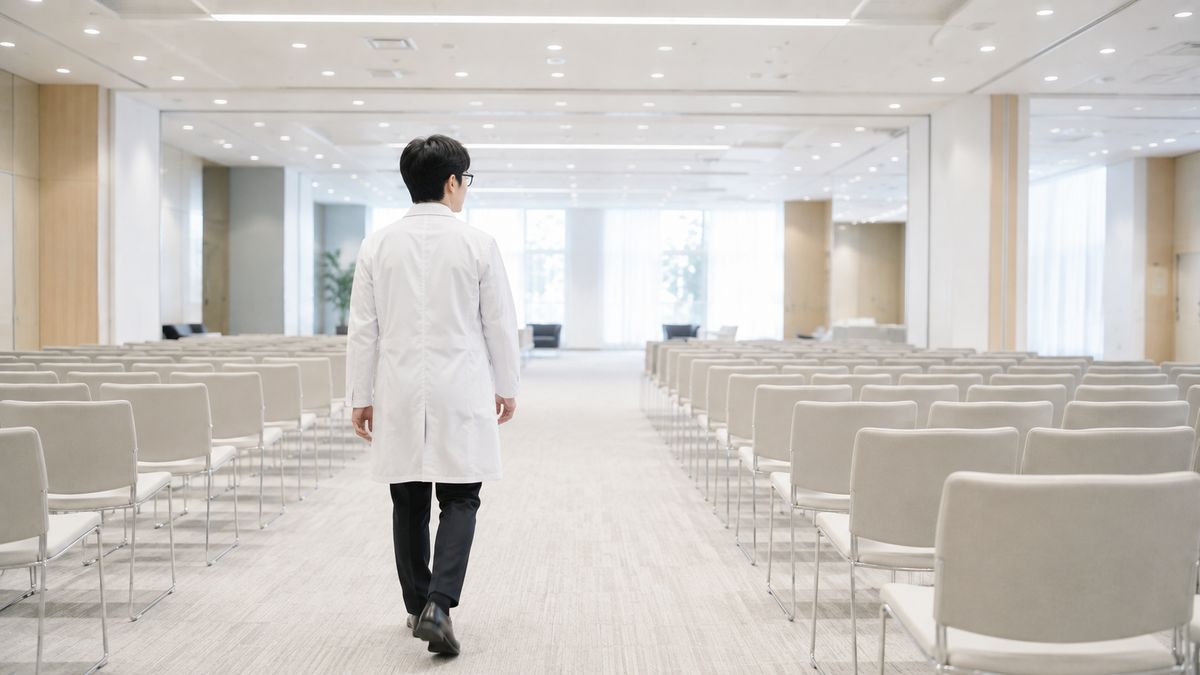 椅子が並ぶ空間を歩く白衣の男性医師の後ろ姿