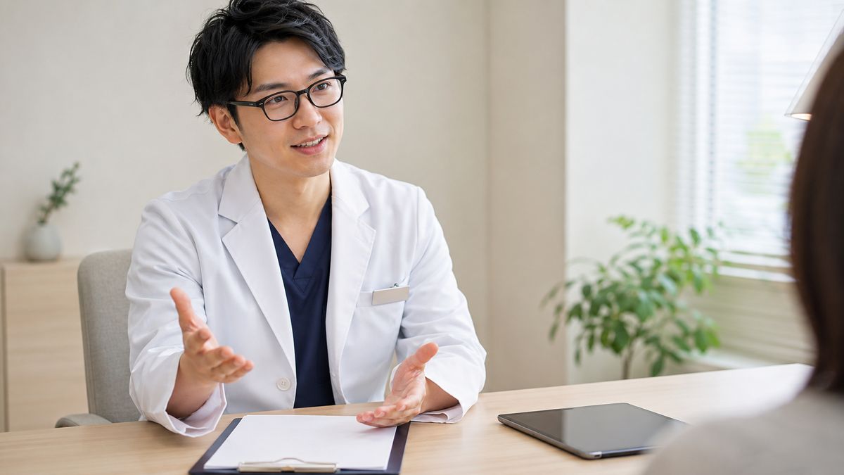 デスクで手ぶりを交えて話す眼鏡の男性医師