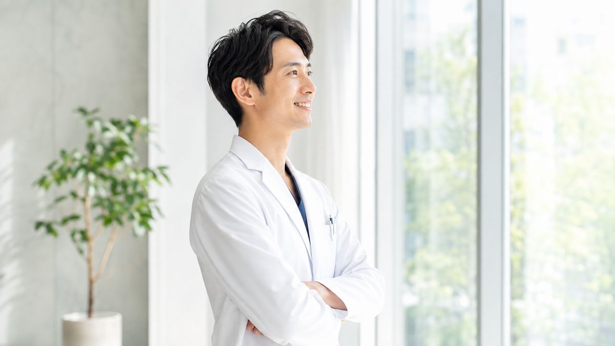 窓辺に立って外を眺める白衣姿の男性医師