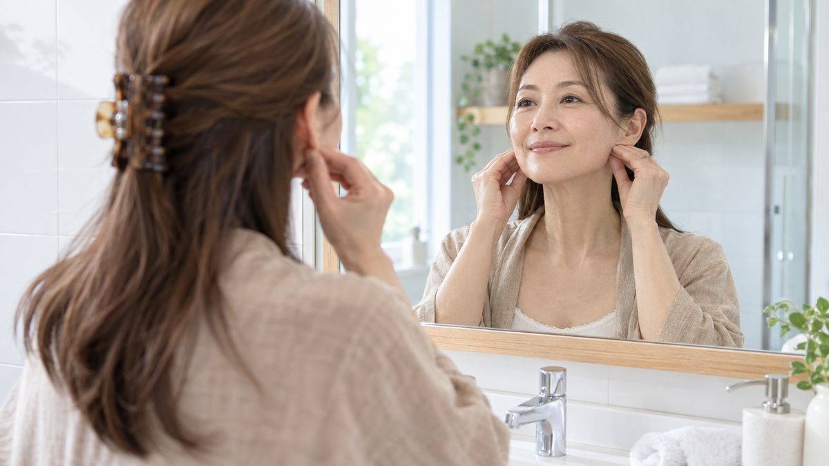 洗面台の鏡の前で耳の前の皮膚を指先で持ち上げる女性